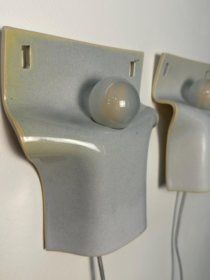 May include: Two wall-mounted ceramic light fixtures featuring a smooth, pale blue-grey glazed finish with subtle yellow-tinted edges. Each lamp has a curved, rectangular body with two rectangular cutouts at the top and a single round glass bulb.