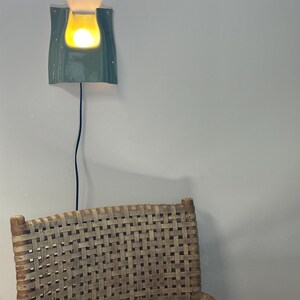 May include: A wall-mounted ceramic sconce with a glossy sage green finish and a curved, U-shaped cutout. The fixture emits a warm, glowing light from a central bulb and features a thin black power cord hanging vertically down the wall.