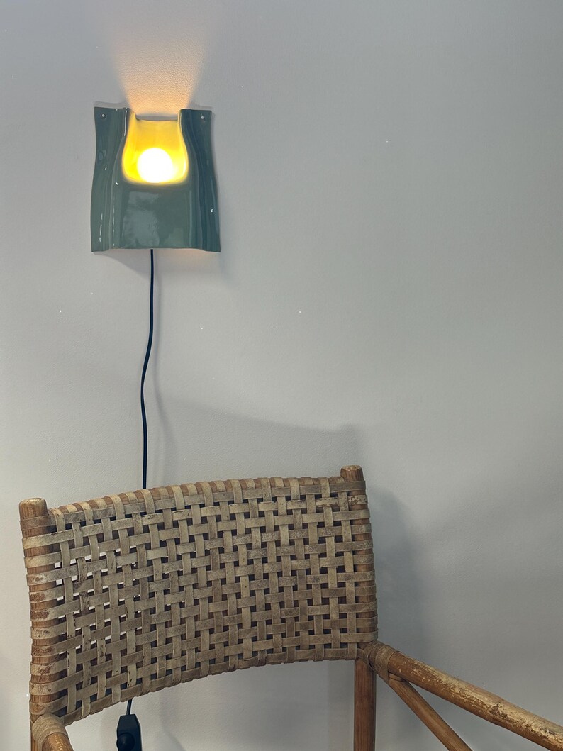 May include: A wall-mounted ceramic sconce with a glossy sage green finish and a curved, U-shaped cutout. The fixture emits a warm, glowing light from a central bulb and features a thin black power cord hanging vertically down the wall.