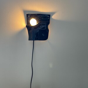 Puede incluir: Un aplique de pared de cer&aacute;mica azul marino oscuro con acabado brillante y forma irregular y plegada. La l&aacute;mpara cuenta con una bombilla redonda de tono c&aacute;lido que sobresale de una abertura en la parte superior izquierda, con un cable negro.