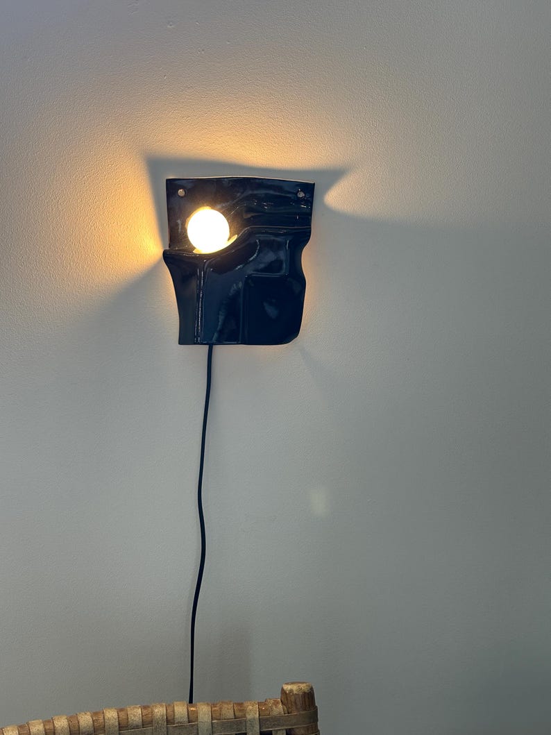 Puede incluir: Un aplique de pared de cer&aacute;mica azul marino oscuro con acabado brillante y forma irregular y plegada. La l&aacute;mpara cuenta con una bombilla redonda de tono c&aacute;lido que sobresale de una abertura en la parte superior izquierda, con un cable negro.