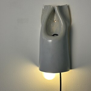 May include: A wall-mounted light fixture with a smooth, glossy, light gray ceramic shade. The shade has a folded, organic shape with a central opening and a small hole for mounting. A single glowing light bulb extends from the bottom.