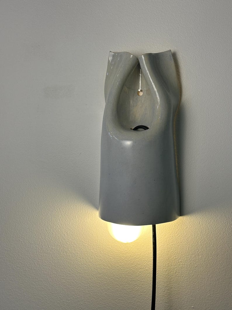 May include: A wall-mounted light fixture with a smooth, glossy, light gray ceramic shade. The shade has a folded, organic shape with a central opening and a small hole for mounting. A single glowing light bulb extends from the bottom.