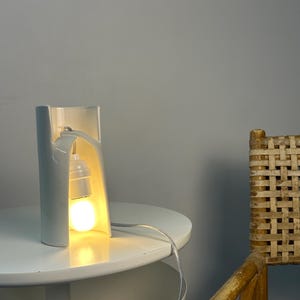 May include: A white ceramic table lamp with a curved, open-front cylindrical structure. The lamp features a single light bulb suspended within the hollow center, emitting a warm glow. A thin, clear electrical cord extends from the base.