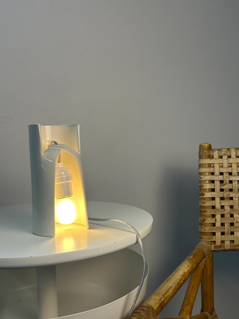 May include: A white ceramic table lamp with a curved, open-front cylindrical structure. The lamp features a single light bulb suspended within the hollow center, emitting a warm glow. A thin, clear electrical cord extends from the base.