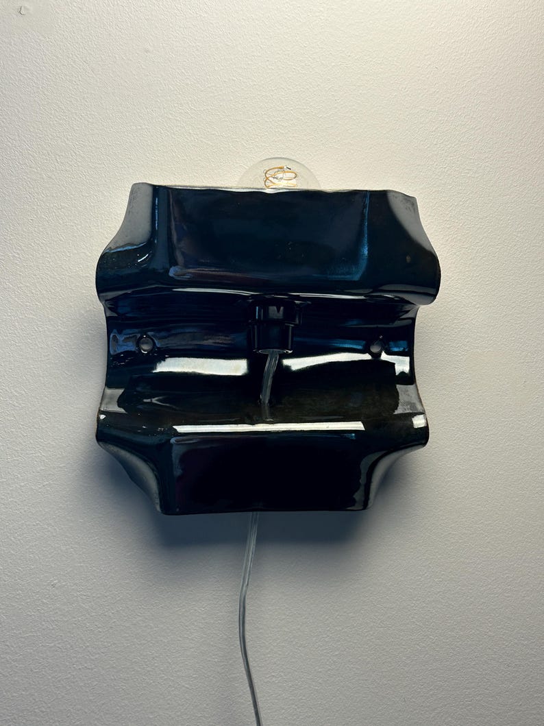 May include: A glossy black ceramic wall sconce with a curved, undulating form. The fixture features a central socket holding a clear glass light bulb with a visible filament. A thin, transparent electrical cord extends downward from the base.