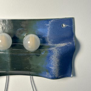 May include: A rectangular wall-mounted ceramic light fixture with a wavy, curved edge. The surface features a blended glaze of deep forest green and vibrant cobalt blue with a textured, speckled finish. Two round, frosted white glass light bulbs are attached.
