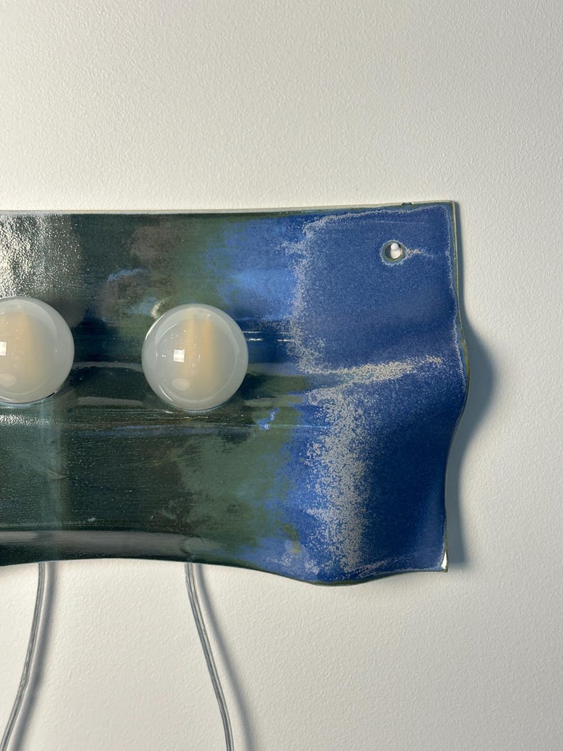 May include: A rectangular wall-mounted ceramic light fixture with a wavy, curved edge. The surface features a blended glaze of deep forest green and vibrant cobalt blue with a textured, speckled finish. Two round, frosted white glass light bulbs are attached.