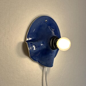 May include: A unique, blue ceramic wall sconce with a glossy finish. The fixture features a bulb socket and a glowing light bulb. A silver cord extends from the base. The sconce is mounted on a light gray wall.