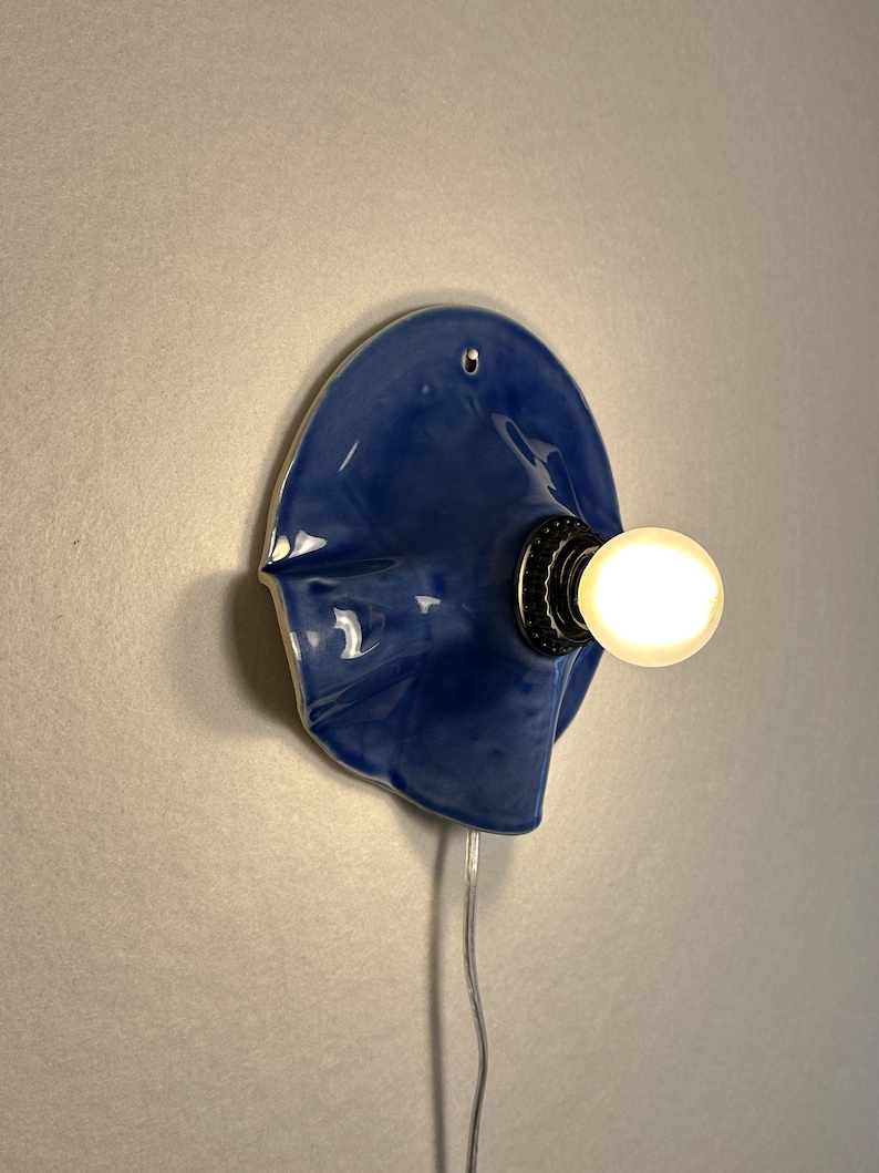 May include: A unique, blue ceramic wall sconce with a glossy finish. The fixture features a bulb socket and a glowing light bulb. A silver cord extends from the base. The sconce is mounted on a light gray wall.