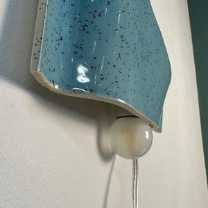 May include: A wall-mounted light fixture featuring a curved, light blue ceramic shade with a speckled dark brown pattern. The fixture includes a single, round, frosted white glass bulb suspended from the bottom, connected by a clear power cord.