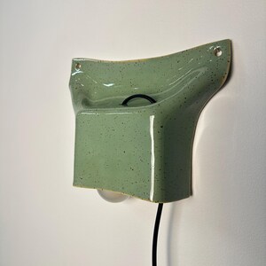 May include: A wall-mounted ceramic light fixture in a light green speckled glaze. The piece features a curved, flared top section with two mounting holes and a rectangular lower housing that holds a single light bulb, connected by a black cord.