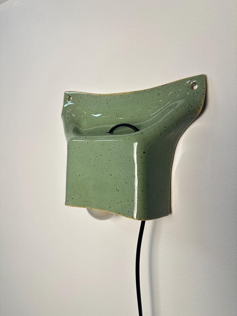 May include: A wall-mounted ceramic light fixture in a light green speckled glaze. The piece features a curved, flared top section with two mounting holes and a rectangular lower housing that holds a single light bulb, connected by a black cord.