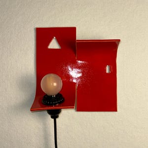 May include: A modern red wall sconce with a glossy finish. The sconce features a geometric design with a triangle cutout and a small rectangular cutout. A spherical light bulb sits on a black base, connected to a black cable.
