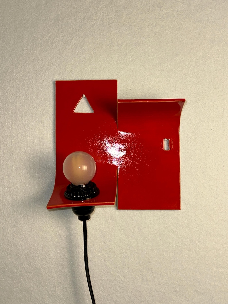 May include: A modern red wall sconce with a glossy finish. The sconce features a geometric design with a triangle cutout and a small rectangular cutout. A spherical light bulb sits on a black base, connected to a black cable.