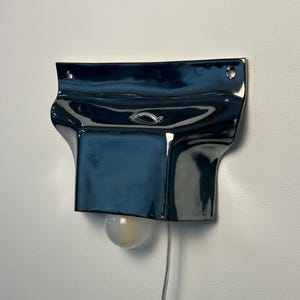 May include: A glossy, dark navy blue ceramic wall sconce with a wide, flared top section featuring two mounting holes. The fixture has a central rectangular body with a small metal loop and a single round, frosted light bulb extending from the bottom.