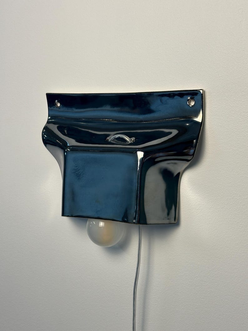 May include: A glossy, dark navy blue ceramic wall sconce with a wide, flared top section featuring two mounting holes. The fixture has a central rectangular body with a small metal loop and a single round, frosted light bulb extending from the bottom.