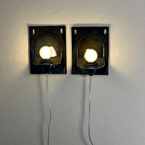 May include: Two wall-mounted sconces with a dark, textured rectangular base and a curved, metallic shade. Each sconce features a small, round light bulb and a black base. The sconces are connected to a wall outlet via a silver wire.