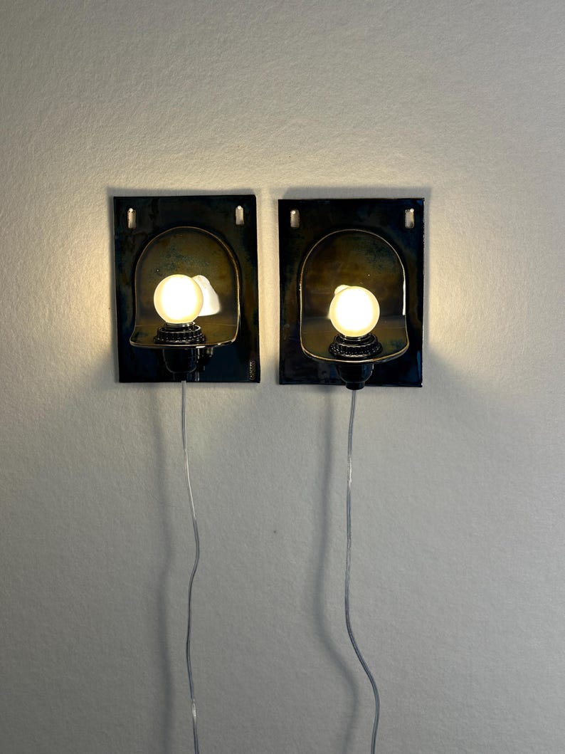 May include: Two wall-mounted sconces with a dark, textured rectangular base and a curved, metallic shade. Each sconce features a small, round light bulb and a black base. The sconces are connected to a wall outlet via a silver wire.