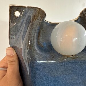 May include: A wall-mounted ceramic light fixture featuring a glossy, mottled blue and brown glaze. The sculptural form includes a circular opening for mounting and a single, spherical, frosted white glass light bulb protruding from the front surface.