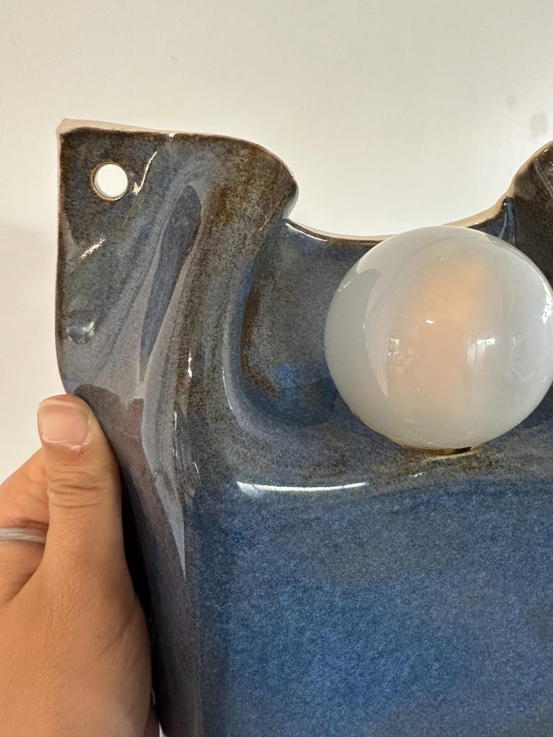 May include: A wall-mounted ceramic light fixture featuring a glossy, mottled blue and brown glaze. The sculptural form includes a circular opening for mounting and a single, spherical, frosted white glass light bulb protruding from the front surface.