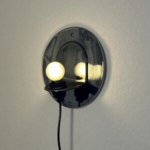 May include: A circular, glossy black ceramic wall sconce featuring a small, glowing spherical light bulb mounted on a protruding shelf. The fixture includes a black power cord hanging downward and a small Taurus zodiac symbol etched at the top.