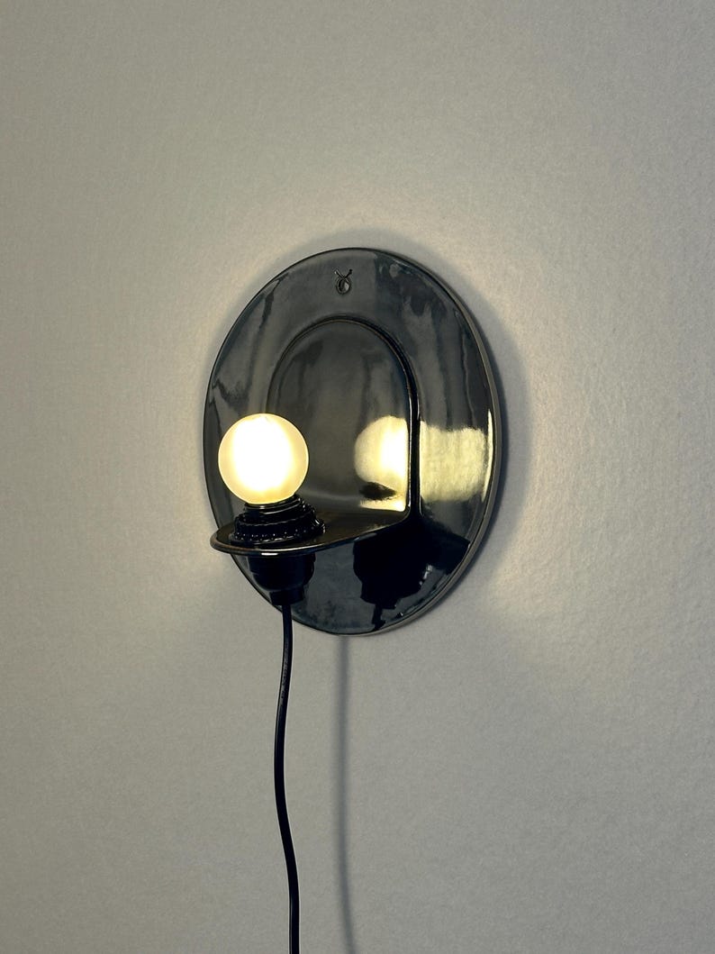May include: A circular, glossy black ceramic wall sconce featuring a small, glowing spherical light bulb mounted on a protruding shelf. The fixture includes a black power cord hanging downward and a small Taurus zodiac symbol etched at the top.