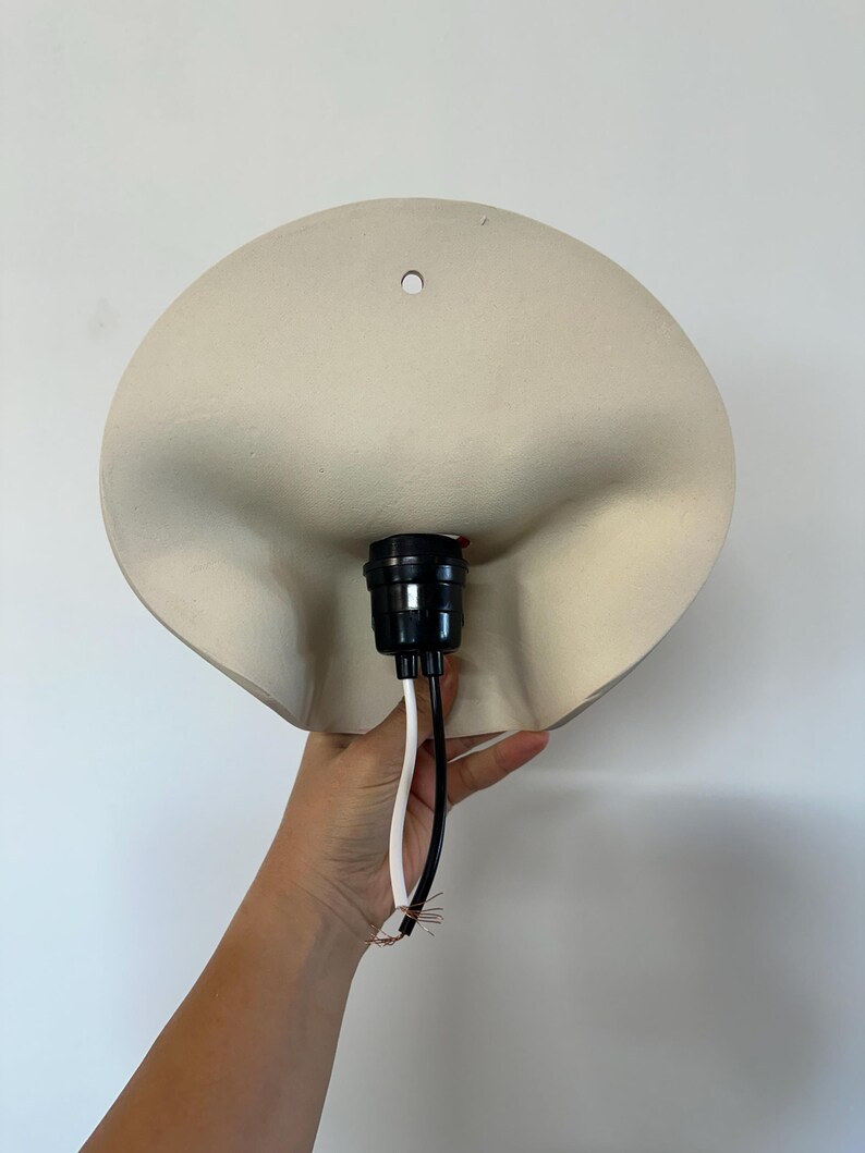 May include: A matte off-white wall lamp fixture with a curved, organic shape and a single mounting hole at the top. The center features a black plastic light socket assembly with one white wire and one black wire extending from the base.