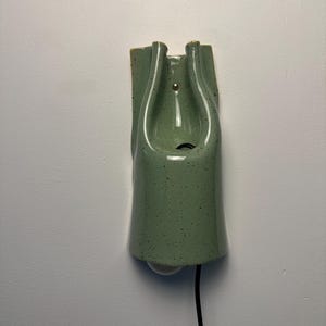 May include: A wall-mounted ceramic light fixture in a sage green glaze with dark speckles. The piece features a smooth, curved, elongated form with a central opening for a light bulb and a black electrical cord extending from the bottom.