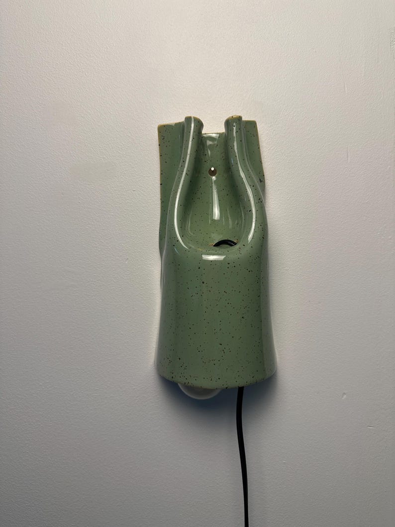 May include: A wall-mounted ceramic light fixture in a sage green glaze with dark speckles. The piece features a smooth, curved, elongated form with a central opening for a light bulb and a black electrical cord extending from the bottom.