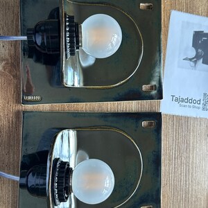 May include: Two dark green ceramic wall sconces with a semi-circular design, each holding a round light bulb. The sconces have a black bulb socket and a small rectangular base. The brand name "Tajaddod" is visible on the base.