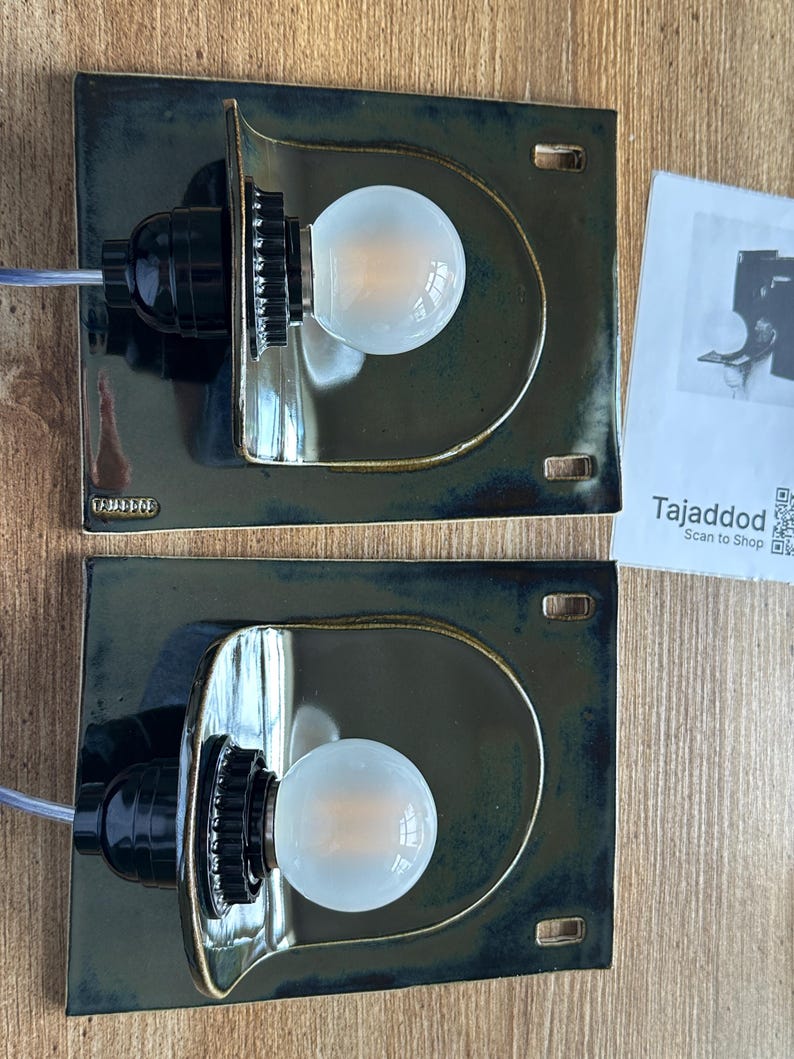 May include: Two dark green ceramic wall sconces with a semi-circular design, each holding a round light bulb. The sconces have a black bulb socket and a small rectangular base. The brand name "Tajaddod" is visible on the base.