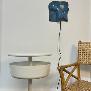 May include: A white metal side table with a circular top and a deep bowl-shaped storage compartment, positioned next to a woven rattan chair and a blue ceramic wall lamp with a single exposed light bulb and a black power cord.