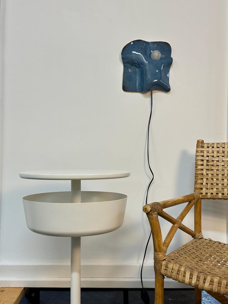 May include: A white metal side table with a circular top and a deep bowl-shaped storage compartment, positioned next to a woven rattan chair and a blue ceramic wall lamp with a single exposed light bulb and a black power cord.