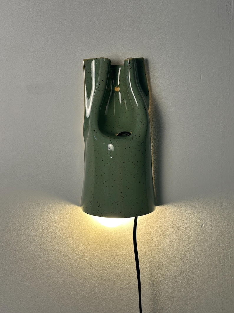May include: A wall-mounted ceramic sconce in a sage green speckled glaze. The fixture features a vertical, folded form with a central opening and a small circular hole near the top. A warm light emits from the bottom, and a black power cord hangs down.