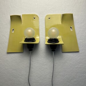 May include: A pair of wall sconces featuring pale yellow, curved ceramic backplates with a glossy finish. Each fixture includes a black socket holding a round, frosted glass light bulb, with a thin gray electrical cord extending downward.