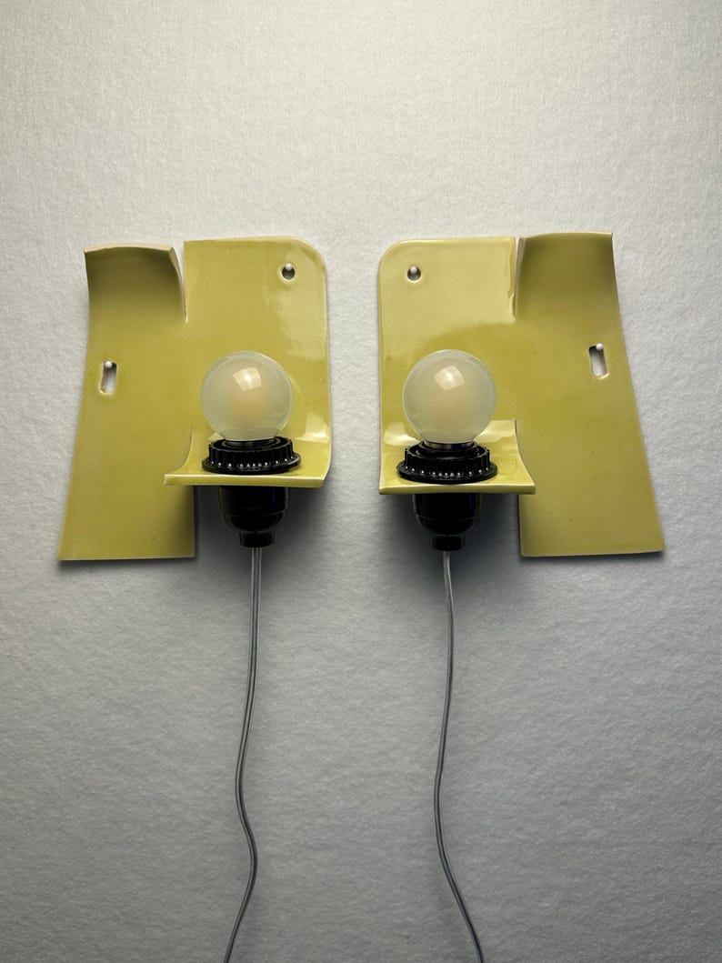 May include: A pair of wall sconces featuring pale yellow, curved ceramic backplates with a glossy finish. Each fixture includes a black socket holding a round, frosted glass light bulb, with a thin gray electrical cord extending downward.