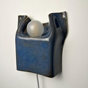 May include: A wall-mounted ceramic lamp with a glossy, deep blue glaze and an organic, curved shape. The fixture features a central circular cutout holding a frosted white spherical light bulb and includes two small metal-rimmed holes for mounting.