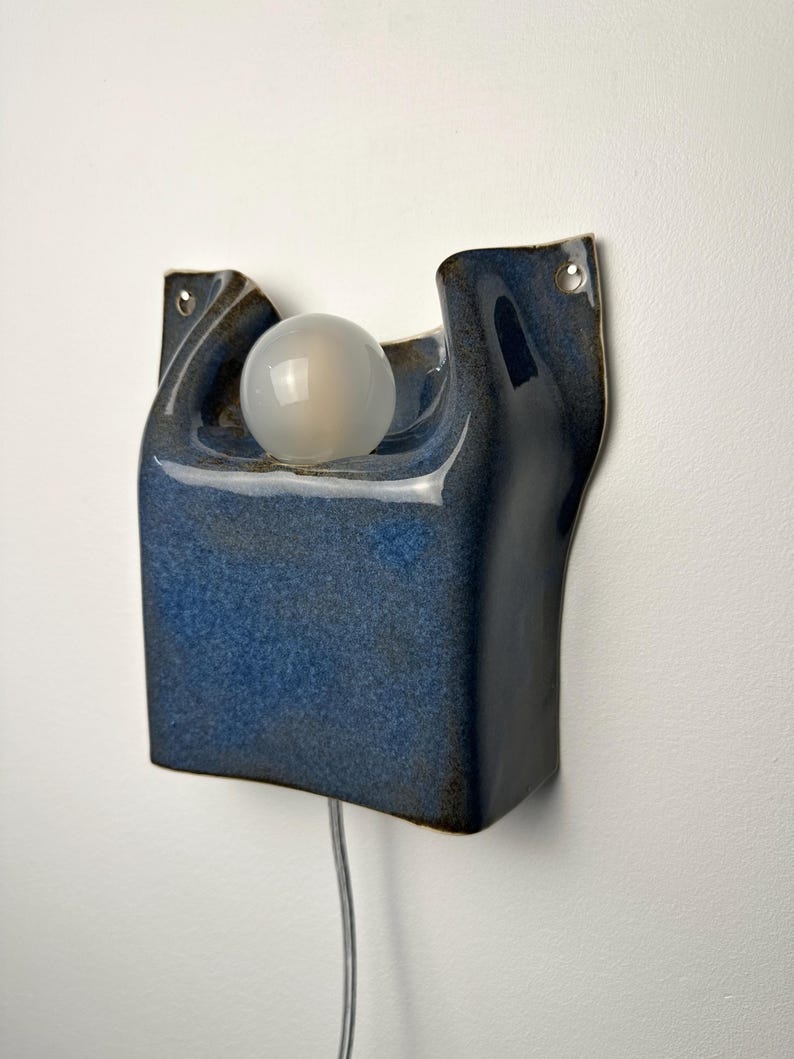 May include: A wall-mounted ceramic lamp with a glossy, deep blue glaze and an organic, curved shape. The fixture features a central circular cutout holding a frosted white spherical light bulb and includes two small metal-rimmed holes for mounting.