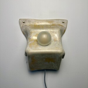 May include: A wall-mounted ceramic light fixture with a mottled beige and cream glaze. The rectangular base features two mounting holes at the top corners and a central circular opening holding a small, spherical, frosted white glass light bulb.