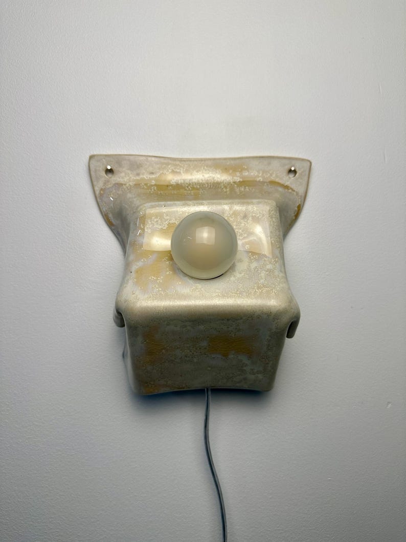 May include: A wall-mounted ceramic light fixture with a mottled beige and cream glaze. The rectangular base features two mounting holes at the top corners and a central circular opening holding a small, spherical, frosted white glass light bulb.