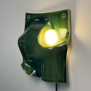 May include: A wall-mounted ceramic light fixture featuring a glossy, deep green glazed surface with an organic, undulating shape. A circular, frosted glass bulb is embedded in the center, emitting a warm, bright glow from the ceramic form.