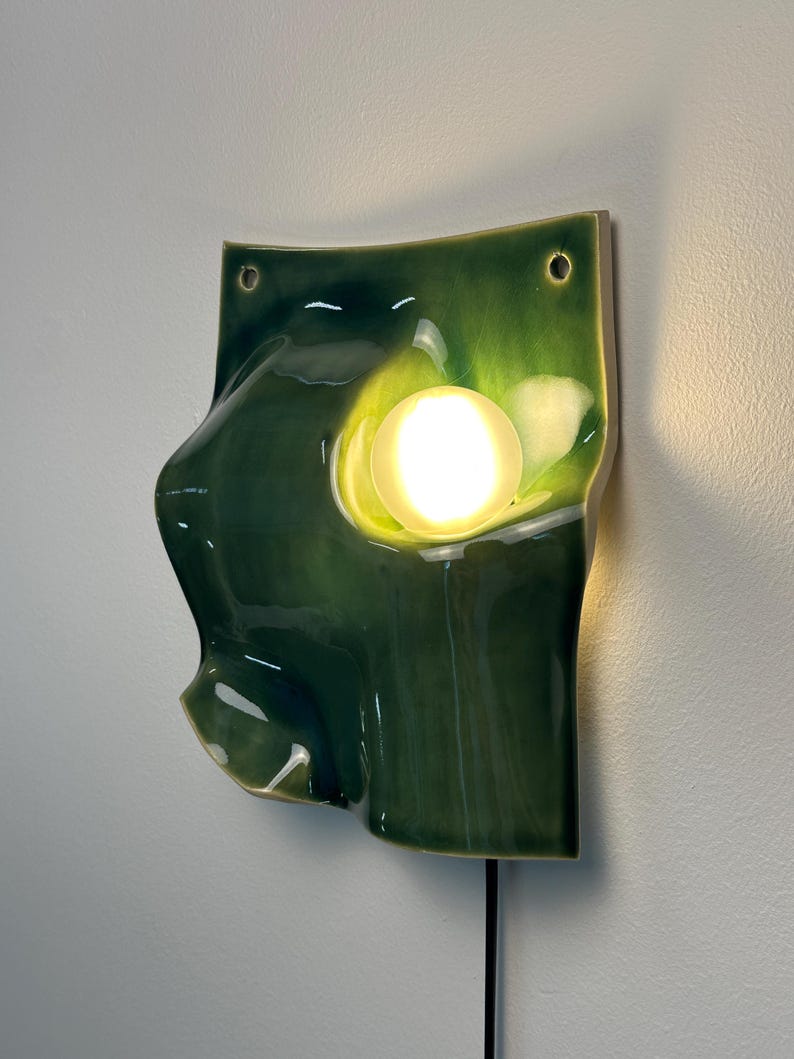 May include: A wall-mounted ceramic light fixture featuring a glossy, deep green glazed surface with an organic, undulating shape. A circular, frosted glass bulb is embedded in the center, emitting a warm, bright glow from the ceramic form.