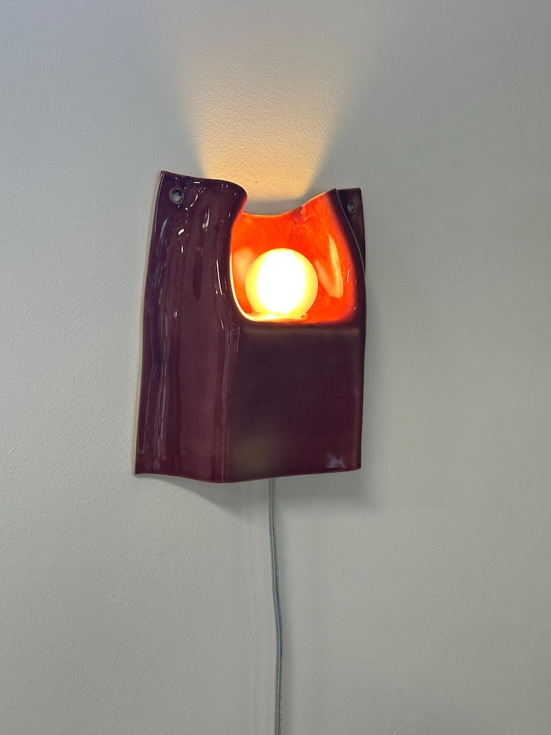 Puede incluir: Un aplique de pared de cer&aacute;mica con un exterior de color ciruela oscuro brillante y un interior naranja vibrante. La pieza presenta un recorte central que alberga una bombilla esf&eacute;rica brillante y dos orificios de montaje en las esquinas.