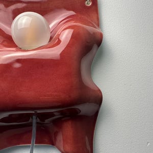 K&ouml;nnte beinhalten: Eine Wandleuchte mit einem gl&auml;nzenden, tiefroten Keramiksockel in wellenf&ouml;rmiger, organischer Form. Eine einzelne, kugelf&ouml;rmige, mattwei&szlig;e Glasgl&uuml;hbirne ist in eine runde Aussparung eingelassen, erg&auml;nzt durch ein transparentes Stromkabel.