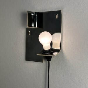 Pode incluir: Uma arandela de parede preta moderna com um design curvo. A arandela apresenta uma l&acirc;mpada branca e um cabo de alimenta&ccedil;&atilde;o preto. A lumin&aacute;ria &eacute; montada em uma parede cinza, criando uma est&eacute;tica minimalista.