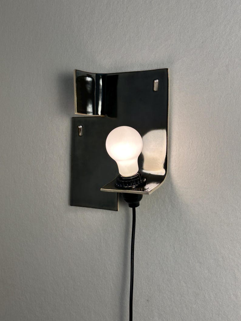 Pode incluir: Uma arandela de parede preta moderna com um design curvo. A arandela apresenta uma l&acirc;mpada branca e um cabo de alimenta&ccedil;&atilde;o preto. A lumin&aacute;ria &eacute; montada em uma parede cinza, criando uma est&eacute;tica minimalista.