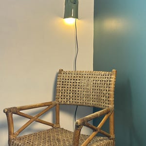 May include: A woven rattan chair with a light brown finish and cross-braced armrests, positioned below a wall-mounted ceramic light fixture with a sage green glaze that emits a warm, glowing light from a single bulb and a thin black cord.