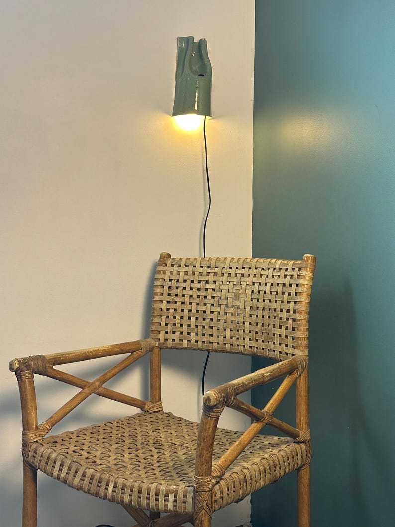May include: A woven rattan chair with a light brown finish and cross-braced armrests, positioned below a wall-mounted ceramic light fixture with a sage green glaze that emits a warm, glowing light from a single bulb and a thin black cord.