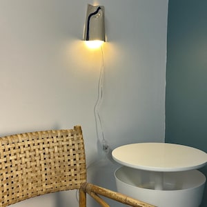 May include: A wall-mounted light fixture with a conical, off-white ceramic shade featuring a single vertical dark blue painted stripe. The lamp emits a warm, downward-facing glow and includes a thin, transparent power cord with a small switch.